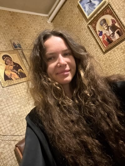 A close-up, eye-level selfie is taken by a woman with long, wavy, dark brown hair. She is wearing a black garment. The background consists of textured, beige wallpaper with a repeating floral pattern. Several framed religious icons are visible on the wall: a large icon of the Virgin Mary and child, a smaller icon of two figures, and a larger icon of a saint holding a book and a cross. A framed clock is also present on the wall above one of the icons. The lighting appears to be artificial and warm. The scene suggests an indoor setting, possibly a room in a home or a religious establishment. The image does not provide information about the city of Turinsk, Russia.