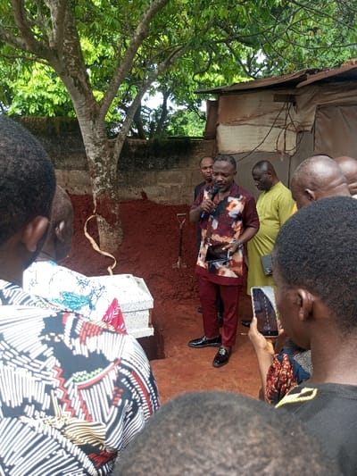 The image depicts an outdoor burial ceremony occurring in Oko Town, Nigeria. A central figure, a man with a beard and mustache dressed in a maroon patterned shirt, maroon trousers, and black patent leather shoes, is speaking into a black microphone. He also holds a dark tablet or smartphone in his left hand. Directly in front of him, to the left, a white coffin with a textured surface, partially covered by red and blue patterned fabric, rests beside an open, rectangular grave dug into reddish-brown soil. A shovel is planted upright in the soil adjacent to the grave.
Attendees surround the scene; in the immediate foreground, the backs of several individuals are visible. One person on the far right holds a white smartphone horizontally, pointed towards the speaker and grave. Another attendee, partially visible to the speaker's right, wears a light olive-green traditional garment. Behind the speaker, a man in a black shirt is visible.
The background features a prominent tree with light-colored bark and green foliage to the left of the grave, backed by a concrete block wall. To the right of the scene, behind the attendees, are rustic structures with corrugated metal roofs and walls, and visible wiring, indicating a residential or community setting. The ground consists of reddish-brown soil.