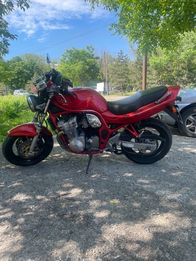 A vibrant red motorcycle is prominently displayed, parked on a gravel surface under bright daylight. The sun casts distinct, dappled shadows from overhead trees onto the ground, indicating it is likely midday or early afternoon.

The motorcycle is a street bike, featuring a black seat, black wheels, and an exposed engine with metallic and chrome components. It has a red front fender, disc brakes on the front wheel, and is supported by a kickstand. Text vertically aligned on the rear fairing reads "CONCAVE IN-LINE FOUR."

The background is filled with lush green trees and dense foliage, suggesting a rural or semi-rural setting. Above the tree line, a clear blue sky with scattered white clouds is visible, crisscrossed by several power lines. A wooden utility pole also stands among the trees. Partially obscured behind the motorcycle's rear wheel is a silver car, and a white van can be faintly seen further in the background. A small, reversed sign visible in the distance appears to spell "ESSOESSE."

The motorcycle is stationary, and no activity or people are present, lending a peaceful atmosphere to the scene in what is identified as Troitskoe, Russia.
