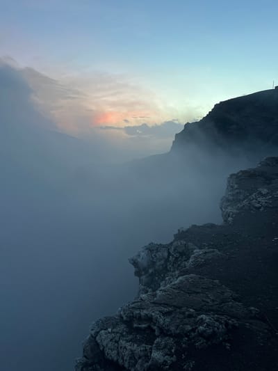 This is a vertical, outdoor, daytime photograph captured from a high vantage point overlooking a misty volcanic landscape. The foreground features a dark, rocky cliff edge with loose, dark soil and stones. The cliff face is rugged and uneven, with jagged rocks and shadows.

In the midground, dense, swirling mist obscures the landscape, creating a sense of depth and mystery. The mist has a bluish-gray hue. Beyond the mist, the silhouettes of more distant mountains or volcanic formations are visible, their shapes softened by the atmospheric conditions.

The sky above transitions from a pale blue at the top to a lighter, almost turquoise hue near the horizon. Wispy clouds are present, with a band of soft pink and orange hues suggesting either dawn or dusk. A small, dark structure, possibly a marker or a building, is perched on the highest point of the furthest visible cliff.

The image does not depict any people, animals, or vehicles. The overall impression is one of natural, rugged beauty and the ethereal quality of mist at an altitude. The scene is likely a volcano crater or a high mountain peak. The reference to Nindirí, Nicaragua, suggests this location is likely Masaya Volcano National Park, known for its volcanic activity and crater lakes often shrouded in mist.