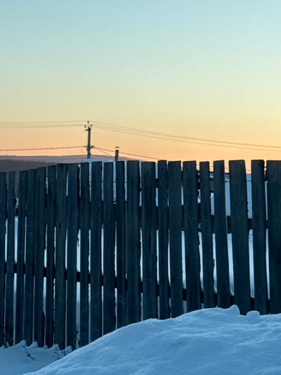 **Location:** Bilimbay, Russia.

**Scene Description:** A forensic view captures a winter landscape under low light, likely dawn or dusk. The image is dominated by a dark wooden picket fence and snow in the foreground, with distant structures and an atmospheric sky in the background.

**Foreground:** A substantial, undulating snowdrift occupies the lower portion of the image, rising from the bottom edge and partially obscuring the base of the fence. The snow is uniformly white with subtle shadows indicating its contours.

**Midground:** A weathered wooden picket fence extends horizontally across the central frame. The vertical pickets are dark, irregular in height and spacing, and show signs of exposure. Gaps between the pickets offer glimpses of additional snow-covered ground beyond.

**Background:** Above the fence, a low, snow-covered horizon line is visible. Centrally positioned is a dark utility pole, equipped with insulators and supporting multiple horizontal power lines that traverse the upper mid-ground. To the right of the utility pole, a slender, dark, chimney-like structure protrudes slightly above the distant terrain.

**Sky:** The upper half of the image features a clear sky with a distinct gradient, transitioning from a pale blue at the zenith to a soft orange-yellow near the horizon, indicative of early morning or late afternoon light.

**People/Actions/Interactions:** No individuals are present, and no actions or interactions are depicted.

**Objects:** Essential objects include the weathered wooden fence pickets, accumulated snow, a utility pole with associated power lines, and a distant chimney-like structure.

**Environment:** The environment is characteristic of a cold, winter setting, likely rural or semi-rural, characterized by snow cover and stark, natural or utilitarian elements under a tranquil sky.