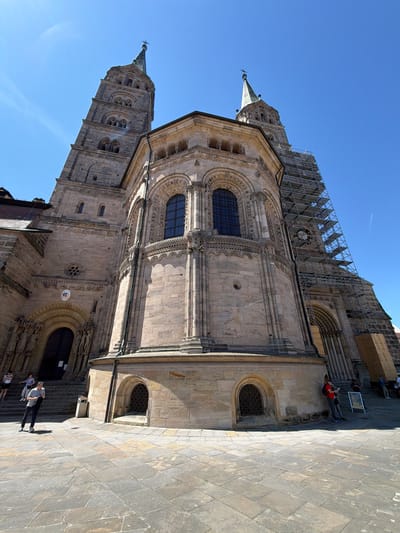 The image shows a low-angle, wide shot of the Bamberg Cathedral in Bamberg, Germany, on a clear, sunny day. The sandstone facade of the cathedral is prominent, with its two imposing towers. One tower on the right is partially obscured by scaffolding. The cathedral features arched windows, intricate carvings, and statues around the main entrance. The foreground is paved with large, irregular stone tiles, and several people are visible in the scene.
On the left, a woman in white shorts and a black top stands on the steps leading up to the entrance. Beside her, a man in a grey t-shirt and dark trousers is looking down at his phone. Further down the steps, another person in white and black is visible.
On the right side of the image, near the base of the cathedral and the scaffolding, a person in a red shirt and dark trousers is leaning against the wall. Another individual in dark clothing stands beside them. A sign with an image is also visible near these individuals. Further in the background, more people are seen on the steps and near the entrance of the cathedral. The sky is a bright, clear blue with a faint contrail from an airplane.