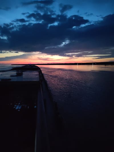 The image shows a dark, twilight scene over a body of water, likely the bay in Kamenniki, Russia. The sky is a deep blue with dramatic, dark clouds illuminated by a faint, vibrant pink and orange sunset on the horizon. A long, dark structure, possibly a pier or breakwater, juts out into the water from the left foreground, its surface appearing somewhat weathered. The water itself reflects the colors of the sky and horizon, with gentle ripples disturbing the surface. In the distance, along the horizon line, faint lights are visible, suggesting a settlement or industrial area. A single, bright blue light emanates from a structure at the far end of the pier. There are no people visible in the image. The overall impression is one of a moody, expansive natural landscape at the close of day.