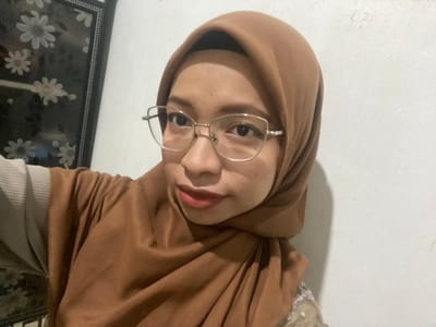 A young woman wearing a brown hijab and glasses is taking a selfie. She is looking directly at the camera with her lips slightly parted. The background is a white wall on the right and a dark decorative panel with floral patterns on the left. The image appears to be taken indoors in a residential setting in Sekar Mawar, Indonesia.
