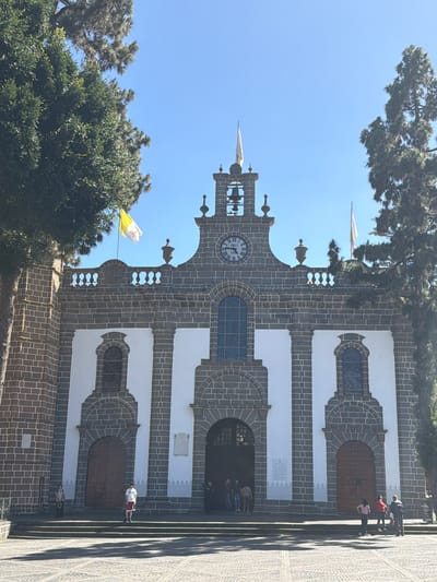 The image shows the exterior of the Basílica de Nuestra Señora del Pino in Teror, Gran Canaria, Spain. The church facade is made of dark volcanic stone and white plaster, with a central arched entrance and two side entrances. Above the main entrance, there is a clock face and a bell tower with two bells. Two flags are visible: a yellow and white flag on the left and a white flag on the right. The church is surrounded by trees and a paved plaza. Several people are visible in front of the church, some standing on the steps and others near the entrances. The sky is clear and blue.