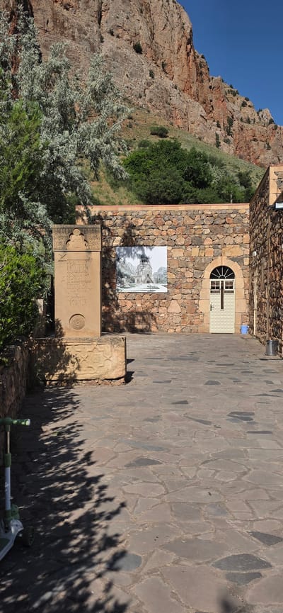 The image shows an outdoor courtyard paved with irregular stone slabs. In the background, a rugged, reddish-brown cliff face rises steeply under a clear blue sky. To the left, a large tree with silvery-green foliage casts shadows on the ground. A stone monument stands to the left of center, featuring engraved text and a decorative carving at the top. Adjacent to the monument is a stone base with geometric carvings. The wall of a building, constructed from rough-hewn stones, runs across the middle ground, displaying a large black and white framed image of a church. To the right of this image, a light-colored door with a distinct cross-shaped handle is set into the stone wall, next to a window with arched panes. Further to the right, another section of the stone building is visible. A small green and white scooter is positioned on the left side of the paved area, partially in shadow. This scene is located in Amaghu, Armenia, likely at the Noravank monastery complex, given the architectural style and the presence of the Momik sculpture.
