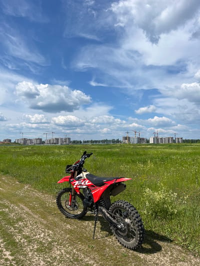 A red and white dirt bike is parked on a dirt path in a grassy field. The motorcycle is angled towards the right, with its front wheel facing left. In the background, a cityscape under construction is visible, with numerous buildings and cranes. The sky is blue with scattered white clouds. The overall scene depicts an outdoor environment on a sunny day, with elements of both nature and urban development. The location is identified as Новочувашский, Russia.