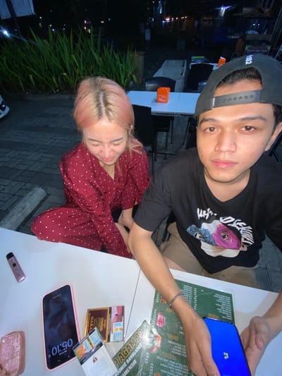 This is a medium shot, taken at night, of a young couple sitting at a white table outdoors. The woman, on the left, has light pink hair and is wearing a dark red shirt with small white polka dots. She is looking down, her eyes are closed, and her mouth is open as if she is speaking or making a sound. The man, on the right, is wearing a black t-shirt with a graphic design on the front and a black baseball cap. He is looking directly at the camera. In front of them on the table are a pink smartphone, a vape pen, two packs of cigarettes, a menu, and a small glittery clutch purse. The background shows some green foliage, tables, and chairs, suggesting an outdoor dining or seating area. The image was taken in Mekar Bakti, Indonesia.