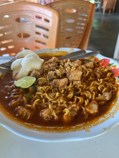 A white, fluted plate prominently displays a dish of instant-style noodles submerged in a dark reddish-brown sauce. The noodles are generously topped with numerous irregular pieces of cooked chicken and three light-colored, crisp crackers. A single green lime wedge is positioned on the left side, adjacent to the crackers. A metal spoon and a second metal utensil are partially submerged within the noodles on the right side of the plate. The plate's upper right rim features a floral design with red and orange elements and green leaves. The dish rests on a white table surface, exhibiting a few small dark specks towards the bottom right. The background is blurred, revealing two light brown plastic chairs with oval-shaped cutouts, suggesting an indoor dining establishment in Sagoe, Indonesia.