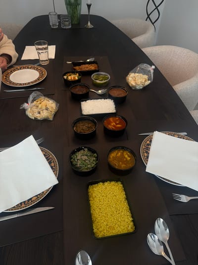 The image depicts a dining table set for a meal, likely in Ridderkerk, Netherlands, as inferred from the context. The table is dark and appears to be made of wood. Several place settings are visible, each with a plate, napkin, and cutlery. The plates have an ornate, gold and black pattern.

Various dishes are arranged on the table, suggesting a meal, possibly Indian or South Asian cuisine. These include containers of rice (both white and yellow), curries or stews in black bowls, a salad or vegetable dish in a black bowl, and a bag of what appear to be puffed snacks or papadums. There are also small containers with a green sauce or relish. A glass of water and two decorative glasses are also present on the table.

In the background, two upholstered chairs are visible, suggesting a home or restaurant setting. A person's arm and shoulder are visible on the left side of the frame, indicating their presence at the table. The overall lighting is somewhat dim, with light sources not directly visible but contributing to a warm ambiance. The scene is static, with no apparent action occurring.