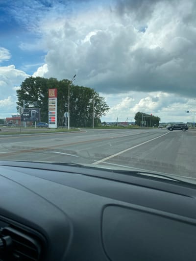 The image shows a view from inside a vehicle, looking out at a roadside scene in Kushnarenkovo, Russia. A Lukoil gas station sign with prices for various fuels is prominently displayed on the right. A billboard advertising coffee is visible on the left. The road ahead is wet, suggesting recent rain, and a few cars are present on the road. Trees line the background, and the sky is filled with dramatic clouds. The scene appears to be a typical view of a gas station and intersection in a rural or suburban area.