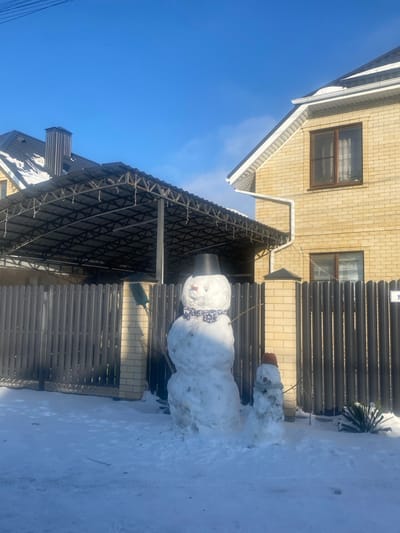A snowman stands in the snow next to a house with a metal fence and a covered area. The snowman has a bucket on its head and a scarf around its middle section. Behind the snowman, a smaller snowman is topped with a flower pot. The sky is blue with light clouds. The house is brick, and another section of the house has windows.