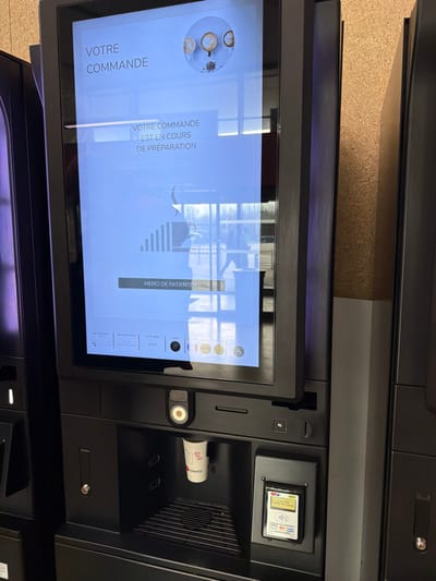 An indoor image captures a black, self-service beverage vending machine in operation, situated within a facility in Toul, France. The prominent vertical touchscreen display shows French text: "VOTRE COMMANDE" (Your Order) at the top, followed by "VOTRE COMMANDE EST EN COURS DE PRÉPARATION" (Your order is being prepared) and "MERCI DE PATIENTER" (Thank you for waiting), along with a progress bar and beverage icons. The screen reflects a bright external environment, revealing windows, bare trees, and the blurred silhouette of a person walking outdoors. Below the screen, a white paper cup bearing "JOY TO GO" and "selecta" branding is positioned in the beverage dispensing bay, above a dark metal drip grate. To the right of the dispensing area, a payment terminal displays "0.00 EUR" and features slots for card insertion, a contactless payment symbol, and logos for Visa and MasterCard. The machine is set against a light brown, possibly wood-paneled wall, with portions of another dark machine visible to the left and a dark panel to the right.