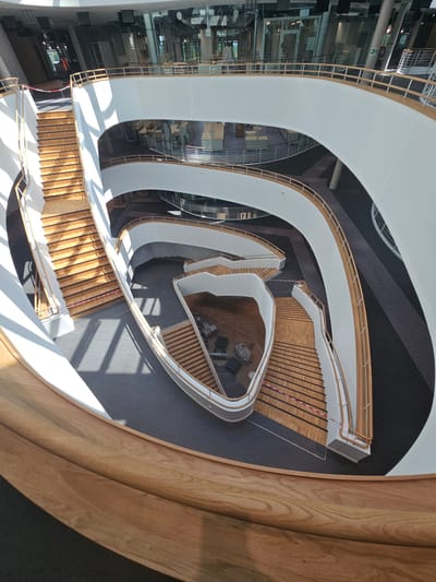 The image shows a multi-story atrium with a large, spiraling wooden staircase. The atrium has white curved walls and railings, with wooden accents on the handrails of the staircase. Sunlight streams in through windows, casting long shadows across the wooden steps. The floor is dark carpet. In the lower left of the image, there are several black cases or containers. In the background, on higher levels, offices or work areas with desks and chairs are visible through glass partitions. The architecture suggests a modern public building, possibly a library, university building, or office complex. The location is specified as Darmstadt, Germany. No people are clearly visible in the image, though a person in an orange vest can be faintly seen on an upper level in one of the cropped hints.