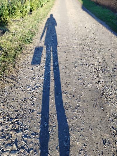The image shows the shadow of a person walking on a dirt road in Vazerki, Russia. The person is carrying a rectangular object, possibly a suitcase or a bag, in their left hand. The shadow is long and stretched, indicating that the sun is low in the sky, likely either early morning or late afternoon. The ground is a mixture of dirt and gravel, with some scattered stones. To the left of the road, there is tall green grass and some dry, straw-like vegetation. A thin, dark line, likely a fence, runs along the right side of the road, with some green grass behind it. The overall scene suggests a rural or semi-rural setting. The time of day, indicated by the long shadow, creates a warm, possibly peaceful atmosphere. There is no visible text in the image.