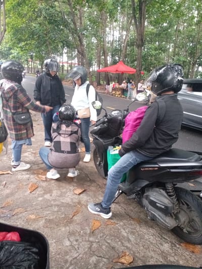 The scene takes place on a paved road in the city of Kecamatan Caringin, Indonesia, at coordinates -7.523 latitude and 107.485 longitude. Five people are standing and interacting on the road. Three of them are wearing motorcycle helmets. A black motorcycle is parked on the side of the road. A man in a black jacket and blue jeans is sitting on the motorcycle. A woman wearing a pink jacket is standing behind the man. Another woman wearing a brown jacket is standing in front of the motorcycle.  A woman wearing a white jacket is standing to the left of the motorcycle. A woman wearing a plaid jacket is standing near the motorcycle.  A red-and-white striped canopy is visible in the background, to the left of the motorcycle.  There are fallen leaves on the ground.  There are trees in the background.  A parked vehicle is visible in the background to the right of the scene.