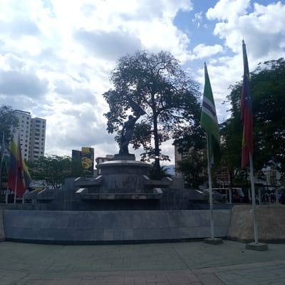The image depicts a monument and surrounding urban environment in El Limón, Venezuela. In the foreground, a paved walkway leads to a multi-tiered concrete monument with a large tree growing behind it. A dark-colored statue of a figure holding a flag stands atop the monument. To the right of the monument, two flagpoles display flags; one is red, yellow, and blue with stars, and the other is green and white striped. In the background, several multi-story buildings are visible, including a tall, light-colored apartment building on the left. A billboard with a yellow background and a dark image is also present. The sky is partly cloudy with patches of blue. The scene appears to be an open public space, possibly a park or plaza, with some people walking in the distance on the right side of the image.