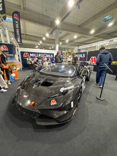 This image captures an indoor motorsport exhibition within a large convention hall in Wolica, Poland. The central subject is a dark grey, carbon fiber-bodied Lamborghini Huracán Super Trofeo Evo racing car, positioned on a dark grey carpeted floor. The car features "MILLERS OILS" branding in red and white, an "OMP" red tow strap on the front, and "motorsport" text on the side fender. Behind the car, several large, golden trophies are displayed on a stand.

Approximately 10-15 individuals are present. In the left foreground, a woman wearing a brown jacket and black top, carrying an orange shoulder bag, stands partially visible. To her right, a person's lower legs in light pants and white sneakers are visible. In the right foreground, a man in a dark blue hooded jacket and blue jeans stands with his back to the camera, observing the car. He is separated from the vehicle by a black stanchion and barrier rope.

In the mid-ground, behind the car, a group of people including a man in a beanie and a person in a camouflage jacket are gathered near a display. A small dirt bike is partially visible behind this group. The background features various exhibition banners and backdrops, prominently displaying "MILLERS OILS" logos in red and white on black and white backgrounds, along with "4T PERFORMANCE" and "RIV" branding. The venue has a high, industrial-style ceiling with visible white metal trussing, exposed ductwork, and numerous bright overhead fluorescent light fixtures.