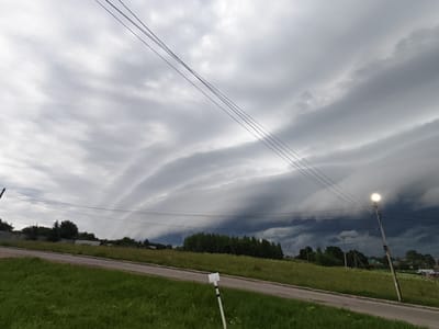 This image depicts a dramatic sky with an approaching storm over a rural landscape in Ateptsevo, Russia. The sky is dominated by heavy, layered clouds with dark, ominous undersides, suggesting imminent rainfall or a thunderstorm. Three thin power lines stretch across the upper portion of the frame, silhouetted against the turbulent sky. In the foreground, a paved road curves to the left, bordered by a grassy embankment. A white sign with black markings is affixed to a pole in the grass. The middle ground features a field of green grass dotted with wildflowers, leading to a dense line of trees. Beyond the trees, a few scattered houses and structures are visible, with a single streetlamp illuminated on the right side of the image. The overall atmosphere is one of impending weather change.
