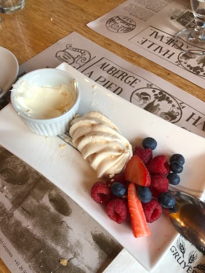 The image shows a close-up, overhead shot of a dessert served on a white rectangular plate. The dessert consists of a meringue, fresh raspberries, blueberries, sliced strawberries, and a small white ramekin filled with a creamy sauce. The plate rests on top of a decorative paper placemat with old-fashioned print. The placemat includes the words "GRUYERES" and "AUBERGE". The placemat mentions "Spécialité fondue Gruyériennes", indicating the dessert is served in the Gruyères region of Switzerland. The text on the placemat, "+41 26 90" hints to a possible phone number. Additional placemats are visible under the main one, one with the text "HALLE AU CHEVAL". Cutlery, including a spoon, is visible above the dessert plate. Part of a glass is also visible in the top left corner. The table surface is made of wood.