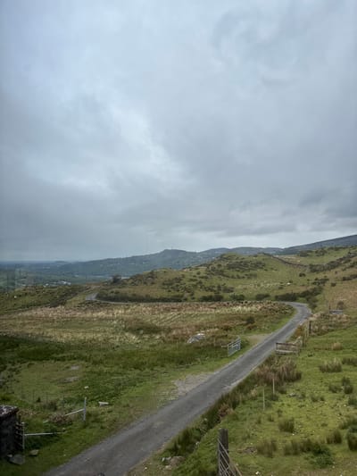 The image presents a landscape in Llanberis, United Kingdom, under a cloudy sky. The scene features a winding, unpaved road cutting through a grassy hillside. The hillside is characterized by a mix of green grass and dry, brown vegetation. Intermittent wooden fence posts and sections of wire fencing are visible along the roadside. The terrain slopes downwards towards a valley where houses or buildings can be partially seen through a haze or fog. The sky is overcast.