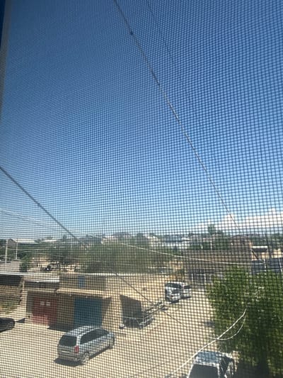 The image shows a view through a window screen of a suburban area. The sky is clear and blue. In the foreground, a silver station wagon is parked on a paved surface. Behind it are several garage structures with different colored doors. Further in the distance, there are buildings and trees. There are power lines visible in the sky. The scene appears to be in Metsamor, Armenia, based on the provided context.