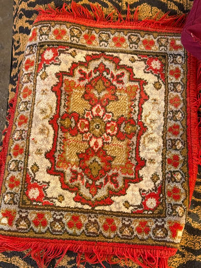 A small, rectangular rug with a red fringe rests on a patterned surface. The rug features a central floral design in red and gold tones against a cream backdrop. A red, patterned border with floral accents surrounds the center. The rug appears textured, with a short pile. The background surface is patterned with an abstract, organic design in shades of brown and gold. A portion of a dark red, soft object is visible in the upper left corner.