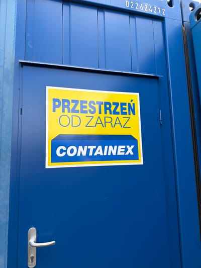 The image shows the side of a blue shipping container. A yellow and blue sticker is affixed to the container door. The sticker has blue text that reads "PRZESTRZEŃ OD ZARAZ" at the top and "CONTAINEX" in a blue box at the bottom. A silver door handle is visible on the lower left side of the door. Above the sticker, a white serial number "022634372" is printed on the container. The container is located in Jodłownik, Poland.