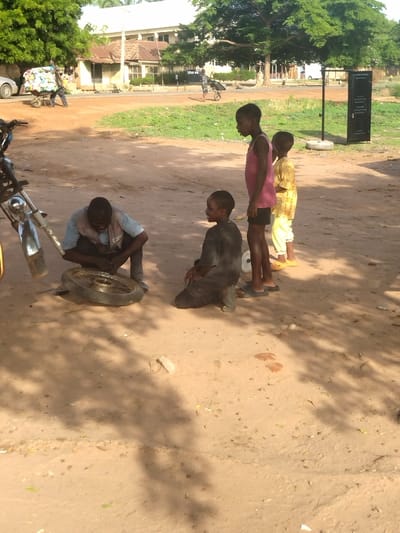 The image depicts a street scene in Kano, Nigeria, at a location with coordinates latitude: 7.683, longitude: 8.537. Four people are present: a man, kneeling and working on a motorcycle tire, a boy kneeling in front of him, and two young girls standing. The man is wearing a blue shirt and a brown vest. The boy is wearing a gray shirt. The girls are wearing pink and yellow shirts. There is a motorcycle lying on its side. The tire is partially removed from the motorcycle. The setting is an open dirt area, with trees and buildings in the background. The people are interacting, likely discussing the motorcycle tire repair. The scene is lit by bright sunlight, casting long shadows from the figures. The surrounding environment is a typical urban setting in Kano, with buildings and trees, indicating a residential or commercial area.