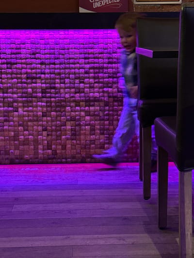 The image captures an atmospheric indoor scene, likely a restaurant or bar, characterized by strong purple and magenta ambient lighting. The main subjects include a young child, decorative architectural elements, and furniture.
A young child is positioned on the left side of the frame, their face and body slightly blurred due to motion, suggesting they are moving or looking around. The child appears to be wearing a light-colored, possibly patterned, top.
The background features a prominent accent wall constructed from numerous small, irregular wooden blocks or tiles, creating a richly textured surface that is vividly illuminated by the purple light. To the right of this wall, a light-colored wooden plank floor extends across the lower portion of the image.
In the upper part of the frame, the dark frames and legs of chairs or stools are visible, suggesting seating arrangements in the establishment. On the far left, a partial sign is present, clearly displaying the word "UNEXPECTED" in white text on a dark background, with other fragmented words above it.
The overall setting implies an evening or nighttime environment, utilizing artificial lighting to create a distinct mood. The child's motion blur adds a dynamic element to the otherwise static interior. The location is specified as Bredene, Belgium.