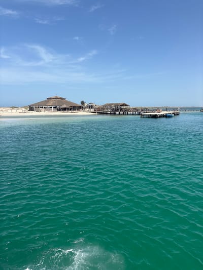 This is a wide-angle, eye-level, outdoor photograph of a tropical island scene from the water. The foreground is dominated by rippling, turquoise water, with a hint of white foam and spray in the lower left corner. In the mid-ground, a sandy island features a large, octagonal thatched-roof building with an open-air structure on stilts to its right. A wooden pier extends from the island, with a small blue boat moored alongside it. The background shows a clear blue sky with scattered white clouds, and a distant hazy horizon. The location is likely on an island off the coast of Ghezine, Tunisia, given the architectural style and the characteristic blue waters of the region. There are no discernible people in the image.