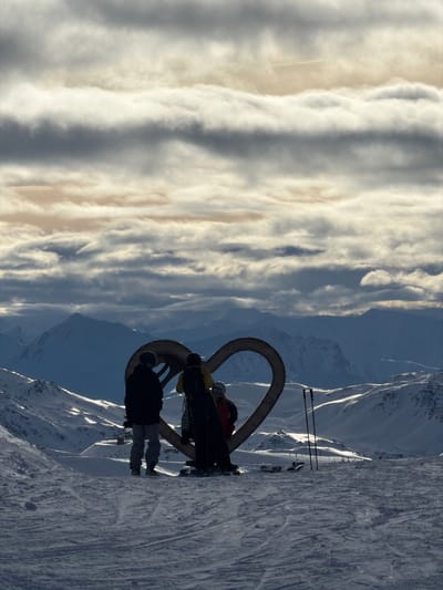 The image presents a breathtaking snowy mountain landscape, rotated 90 degrees clockwise from its natural orientation. When viewed correctly, it depicts a high-altitude scene with a dramatic sky and a group of individuals near a unique art installation.
In the foreground, a vast expanse of snow covers the ground, with visible ski tracks indicating recent activity. Several people, dressed in winter sportswear, are silhouetted against the brighter background. They appear to be skiers or snowboarders, standing near a large, artistic sculpture. This sculpture is a prominent feature, consisting of two intertwined, curved elements, possibly made of wood or metal, forming a looping, abstract design. One person in a dark jacket and light pants stands to the left of the sculpture, while others are partially visible behind and within its curves. A pair of ski poles are stuck upright in the snow, adding to the context of winter sports.
In the mid-ground and background, majestic snow-covered mountains stretch across the horizon. The mountains are rugged and imposing, with some peaks and ridges brightly illuminated by the sun, contrasting with shadowed slopes. The lighting suggests either early morning or late afternoon, creating long shadows and highlighting the natural contours of the landscape.
The sky, dominating the upper portion of the correctly oriented image, is filled with impressive cloud formations. These clouds are elongated and layered, displaying a range of white, grey, and soft golden-beige tones where sunlight catches their edges. The texture and depth of the clouds contribute significantly to the dramatic atmosphere of the scene.
The overall impression is one of grandeur and adventure, capturing a moment of pause during a mountain expedition or ski trip. The combination of the natural beauty of the snowy peaks and dynamic sky with the intriguing man-made sculpture creates a captivating visual narrative.
No visible text is present in the image.