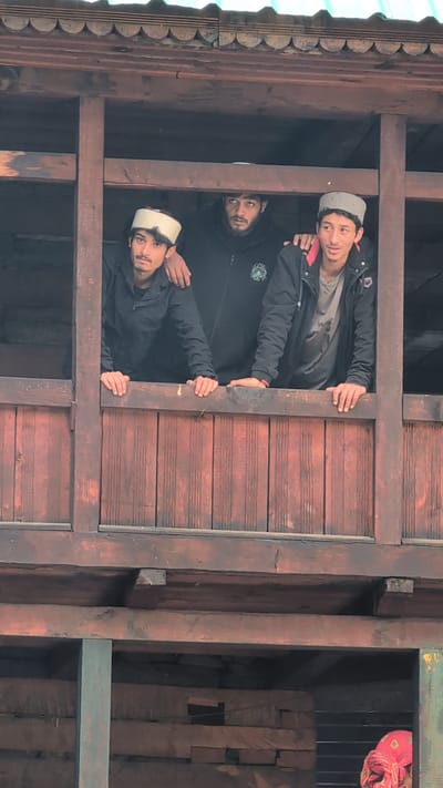Three young men are standing on a wooden balcony in Malana, India. The man on the left wears a white and maroon cap, a black jacket, and is looking at the camera. The man in the middle wears a black hoodie and is looking slightly to the left. The man on the right wears a grey cap, a black jacket over a grey t-shirt, and is looking to his right. The balcony is made of dark wood planks and posts, and the building appears to be constructed from wood as well. In the background, another person wearing a pink headscarf is partially visible. The lighting suggests it is daytime.