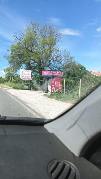 The image, taken from inside a vehicle, shows a roadside scene in Seltsi, Bulgaria. On the right side of the road is a small, red and blue "VENDING" kiosk advertising drinks and snacks. To the left of the kiosk is a gate, and beyond it is a sign advertising construction materials. A green trash bin is situated near the sign. The sky is blue with scattered clouds, and trees are present in the background. A section of the vehicle's dashboard is visible in the foreground.