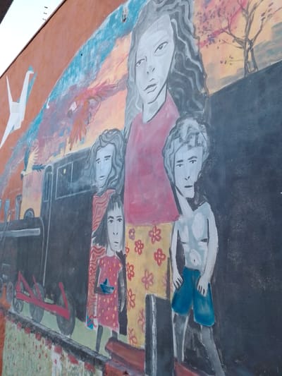 This is a large, colorful mural painted on an outdoor wall, located in Campo Grande, Brazil. The mural depicts a stylized scene featuring multiple human figures and elements of nature and transportation.

The central figure is a female with pale skin, dark eyes, and long, wavy dark hair, wearing a red short-sleeved top and a yellow skirt adorned with red floral patterns. To her immediate left stands another female figure with similar pale skin and dark, wavy hair, dressed in a red and white striped garment. Partially in front of this figure is a smaller girl with straight, dark hair, wearing a red dress with yellow polka dots and holding a small blue origami boat in her hands. To the right of the central female figure, a boy with short, light-colored wavy hair is depicted bare-chested, wearing blue shorts.

Behind the figures and extending to the left, a dark-colored train engine and tender are illustrated on railway tracks. The ground beneath the train and figures is textured with patterns of green and red, suggesting grass or foliage. The upper portion of the mural features a sky transitioning in color from reddish-orange on the left to a darker blue on the right. In the upper left, a white origami crane is shown in flight. Below it, a large, reddish-orange bird with outstretched wings soars. A broad, light blue, abstract band arcs across the upper part of the sky, with a small, dark sailing ship faintly visible within it in the upper right corner. Further to the right, stylized trees with pink or red blossoms are depicted against a yellowish-orange background. The artistic style is illustrative, with distinct outlines and flat color application.