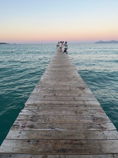 The image depicts a pier stretching out into the ocean, with a group of people gathered at the end of it. The pier is constructed from weathered wooden planks. To the left of the pier, the sky transitions through a gradient of colors, from a pale blue near the horizon to soft hues of pink and yellow, suggesting either sunrise or sunset. A faint rainbow can be observed in this colorful sky. The ocean is a deep teal color, with visible ripples on its surface, indicating a gentle breeze or movement of water. In the distance, a mountain range is faintly visible on the horizon. Several buoys are scattered in the water. The people on the pier appear to be dressed in casual attire. Their postures suggest they are observing the scenery or interacting with each other. The overall mood is serene and peaceful, evoking a sense of calm and natural beauty. There is no visible text in the image. The scene is likely set in Platja De Muro, Spain.