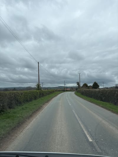 The image depicts a rural road in Longden, United Kingdom, under an overcast sky. The road is asphalt, with a white line marking the center. Hedges and grass line both sides of the road. Utility poles and wires run along the left side of the road. The view suggests a perspective from inside a vehicle.