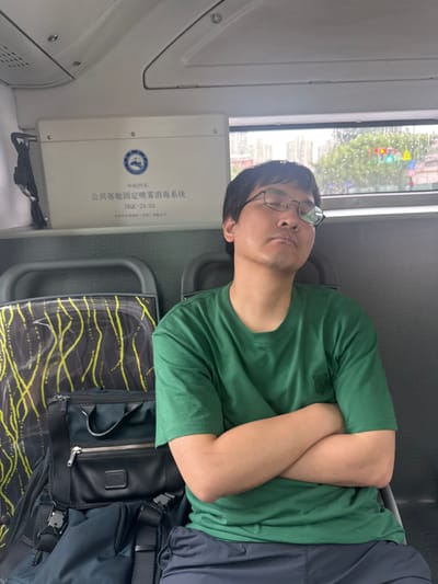 The image shows a man wearing glasses and a green t-shirt sleeping with his arms crossed while sitting on a bus. He is seated next to luggage. The interior of the bus appears to be modern with overhead compartments and a window showing an urban landscape with trees and traffic lights. The text on the overhead compartment suggests the bus is part of public transportation in Xuhui District, China, likely related to a disinfection system.