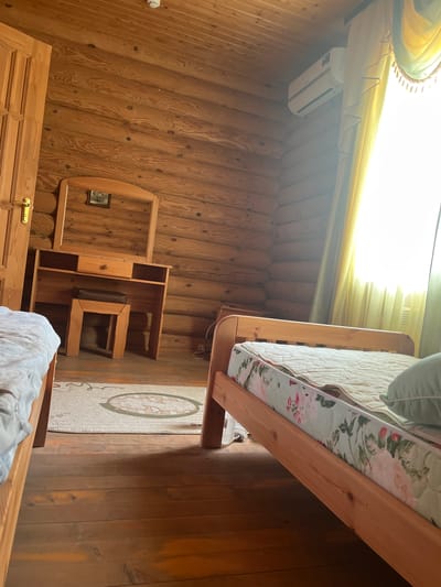 The image shows a bedroom in a log cabin. The walls and ceiling are made of light-colored logs. A wooden door with a brass handle is visible on the left. In the center of the room is a wooden vanity with a mirror and a small stool. The vanity has drawers and open shelves. A bed with a floral-patterned mattress is on the right side of the room, and another bed is partially visible on the left. A rug with a circular design is on the floor between the vanity and the bed on the right. An air conditioning unit is mounted on the ceiling on the right side of the room. Yellow and green curtains are drawn across a window on the right. The floor is made of wooden planks. The scene depicts an interior of a room, likely a bedroom, in Koshara, Russia.