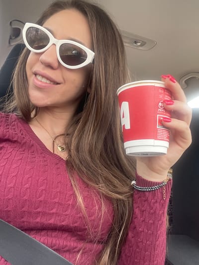 A woman with long brown hair and white sunglasses is seated in a car. She is wearing a burgundy sweater and a gold necklace with a heart-shaped pendant. She holds a red and white paper cup in her right hand, displaying her red nail polish. A black beaded bracelet is on her wrist. A seatbelt crosses her torso. The car interior and visor are visible in the background. The exact location in Iastrebino, Bulgaria, cannot be determined from the image.