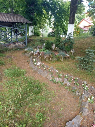 The image depicts a tranquil garden scene with a focus on a decorative arrangement of garden figures resembling geese and ducklings, strategically placed among rocks and small plants. The figures are arranged in a slightly elevated area, creating a focal point within the garden. A path, outlined by rocks, winds through the scene.
To the left of the frame, a partially visible gazebo with a corrugated metal roof stands. The gazebo is constructed with grey metal latticework, and inside, what appears to be a simple structure or perhaps a well is visible. Surrounding the gazebo and extending into the background are mature trees with lush green foliage. Some tree trunks have been painted white near their bases.
In the background, a wooden house with a pink-tiled roof is visible through the trees. The environment suggests a rural or suburban setting, possibly a private garden or a public park area. The ground is covered with a mix of grass, loose soil, and small pebbles. The overall impression is one of a well-maintained, slightly rustic outdoor space.
This specific scene is located in the city of Giaginskaya, Russia. The presence of the gazebo, the style of the house, and the type of vegetation are consistent with typical landscaping and architecture found in this region. The garden appears to be a place of leisure and decoration, designed to be visually appealing and serene.