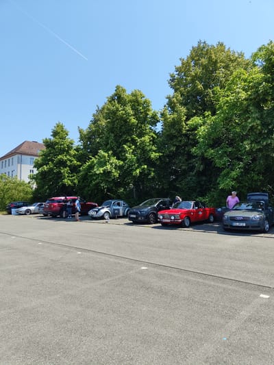 A group of people stand in a parking lot next to several cars, with a building and trees in the background. The cars include a red convertible, a gray hatchback, a silver coupe, and a white sports car. The building is multi-story and appears to be an office or residential building. The trees are tall and leafy, suggesting a warm weather season. The sky is clear and blue, with a faint contrail from an airplane visible. The scene appears to be a casual gathering or event. The license plate on the gray hatchback reads "MZG PR 12", which is a German license plate, and indicates the location is likely Germany. Pirmasens is a city in Germany.