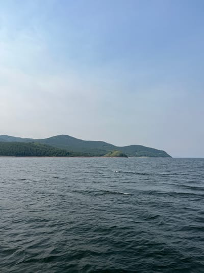 The image shows a wide body of dark water with moderate ripples, stretching towards a distant shoreline. The shoreline is characterized by rolling green hills covered in dense forest. The sky above is a pale blue with hazy clouds. The overall scene depicts a natural landscape, likely a lake or a wide river, given the calm, expansive water and the forested hills. The location is described as being in the city of Novoselovo, Russia, suggesting it is a view from the Krasnoyarskoye Reservoir or a similar body of water within the region. There are no people or man-made structures immediately visible in the frame.