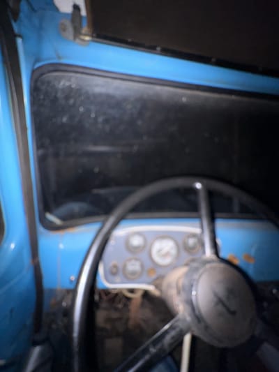 The image shows the interior of an old, blue vehicle, likely a truck or a tractor, with a focus on the dashboard and steering wheel. The dashboard features several circular gauges, some of which are blurry but appear to have needles indicating readings. The steering wheel is black and appears worn. The interior of the vehicle seems somewhat dilapidated, with visible rust on the blue paintwork near the window frame. The overall lighting is dim, suggesting it might be nighttime or the interior is poorly lit. The environment outside the vehicle is dark and indistinct. There are no people visible in the image. The location of Kechut, Armenia, is not discernible from the visual information presented in the image.