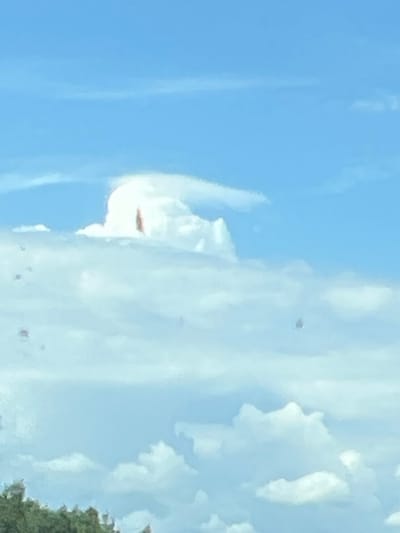 The image depicts a wide shot of a sky filled with clouds. The sky is a vibrant blue, and a variety of cloud formations are visible, ranging from wispy cirrus clouds high up to more substantial cumulus clouds lower down. There are some scattered dark spots within the clouds, which could be debris or lens artifacts. In the distance, a line of green trees is visible at the bottom of the frame. The overall scene appears to be an outdoor environment under a bright, possibly sunny, sky. No specific location within Крупскі сельскі Савет, Belarus can be determined from the image.
