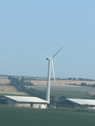 A bright, clear day is depicted in this image, set in Palauzovo, Bulgaria. The scene features a tall, white wind turbine with three blades, its rotor positioned to the upper right. In the foreground, two low-slung, rectangular buildings with white roofs are visible. The building on the left appears to be an industrial or agricultural structure with a light-colored facade and dark openings. To the right, a similar building is seen, with the right half of its roof extending further. Beyond the turbine, a landscape of rolling hills and fields unfolds, with patches of farmland in various shades of brown and green. Dense clusters of trees are scattered across the terrain, with a particularly large, dark mass of foliage to the left of the turbine. The sky is a pale, uniform blue, devoid of clouds.