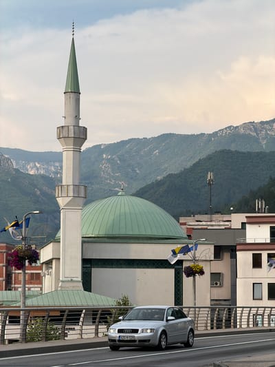 This is a photograph of a mosque with a tall, slender minaret topped with a green spire and a crescent moon detail. The mosque has a large, domed roof, also green. The building appears to be modern, with clean lines and white facade elements. In the foreground, a silver Audi A4 sedan is parked on a street next to the mosque. The car has the license plate "A25-M-528".
The scene is set against a backdrop of lush green mountains. The sky is overcast with some patches of brighter light, suggesting it might be early morning or late afternoon. The overall atmosphere is calm and serene. There are flags flying from a lamppost near the mosque, one of which appears to be the flag of Bosnia and Herzegovina. The location is identified as Konjic, Bosnia and Herzegovina. The photograph is taken from a slightly elevated perspective, possibly from another building or a higher vantage point across the street. No people are visible in the image.