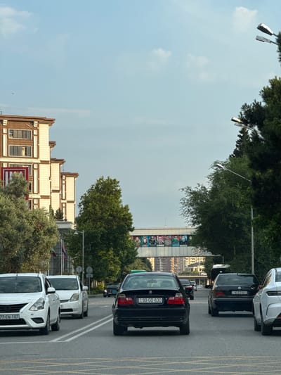 The image depicts a street scene in Baku, Azerbaijan. Several cars are driving on a multi-lane road. In the foreground, a white Nissan sedan with license plate "77-EN-187" is in the left lane. Behind it is another white sedan. In the right lane, a black BMW sedan with license plate "99-GO-830" is visible, followed by a black Mercedes-Benz sedan with license plate "90-EG-601". Further in the background, other vehicles are seen. On the left side of the street, a modern building with a light brown facade and multiple windows stands tall. Trees line the right side of the road. Overhead, a pedestrian bridge is adorned with portraits of several individuals. Streetlights are also present, with two prominently visible in the upper right portion of the image. The sky is partly cloudy.