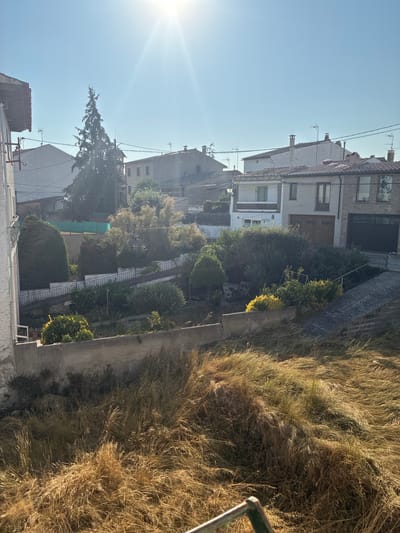 The image depicts a residential area under a clear, sunny sky, with the sun's rays creating a lens flare effect. In the foreground, dry, tall grass covers a slope. A concrete retaining wall runs horizontally across the mid-ground. Behind the wall, a terraced garden with various shrubs and trees is visible. A set of stairs with a black metal railing descends through the garden. In the background, several houses with tiled roofs are clustered together. A tall evergreen tree stands prominently among them. The architecture suggests a European setting, potentially in the town of Castellterçol, Spain, characterized by its traditional residential buildings and natural landscaping. No people or specific actions are captured in the image.