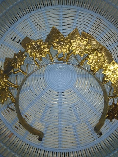 An ornate, golden-toned metallic headpiece is depicted resting on a light blue, woven plastic surface. The headpiece consists of a silver-colored, U-shaped band serving as the base. Attached to this band are multiple upward-projecting golden decorative elements. These elements include stylized, multi-petal floral designs, some featuring small, reflective centers, and triangular plates. Both types of elements are affixed to the base band by thin, coiled golden wires. The woven surface exhibits concentric circular patterns and appears to be part of a basket or mat. There are no individuals, active interactions, or contextual elements to determine a specific location within Ottignies-Louvain-la-Neuve, Belgium, from the image alone.